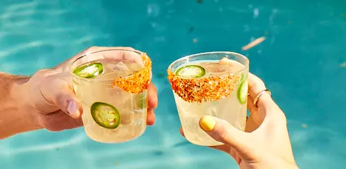 Two hands clink glasses of a clear cocktail with jalapeño slices and chili powder on the rim, held over a blue swimming pool.
