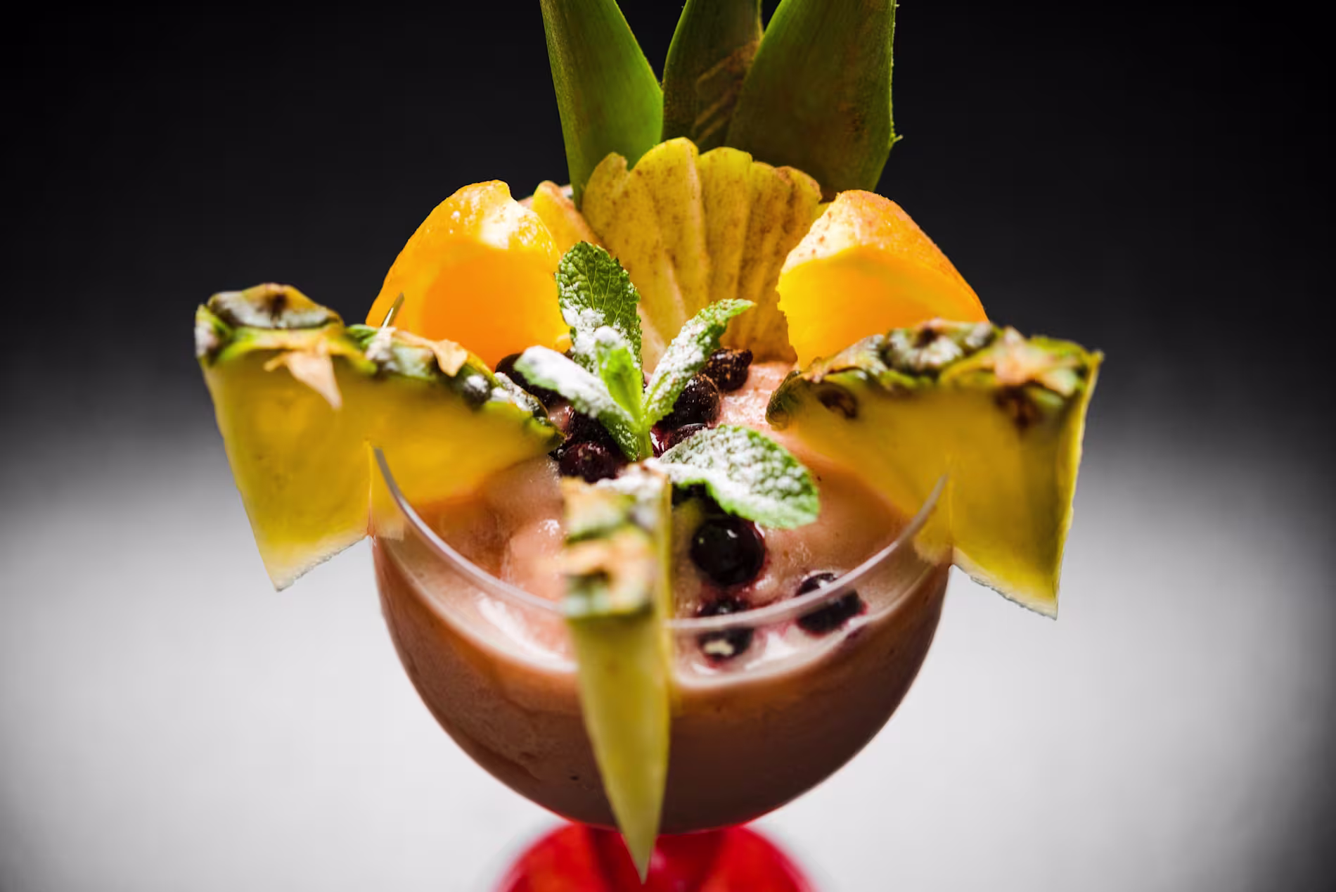 A glass dessert topped with pineapple slices, orange wedges, mint leaves, and powdered sugar, garnished with dark berries.