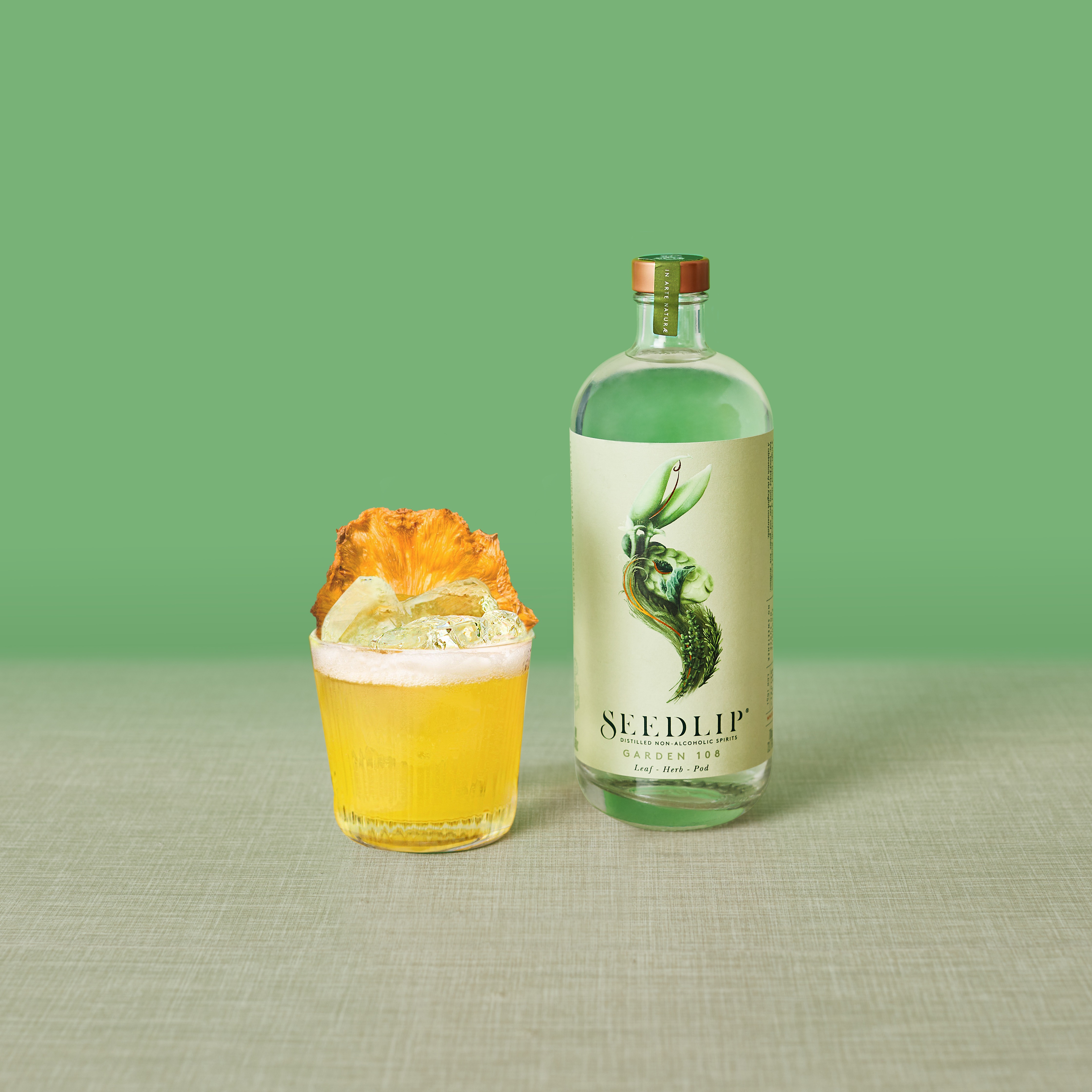 A jalapeno and pineapple margarita in a rocks glass with a dried pineapple garnish, next to a bottle of Seedlip Garden 108.