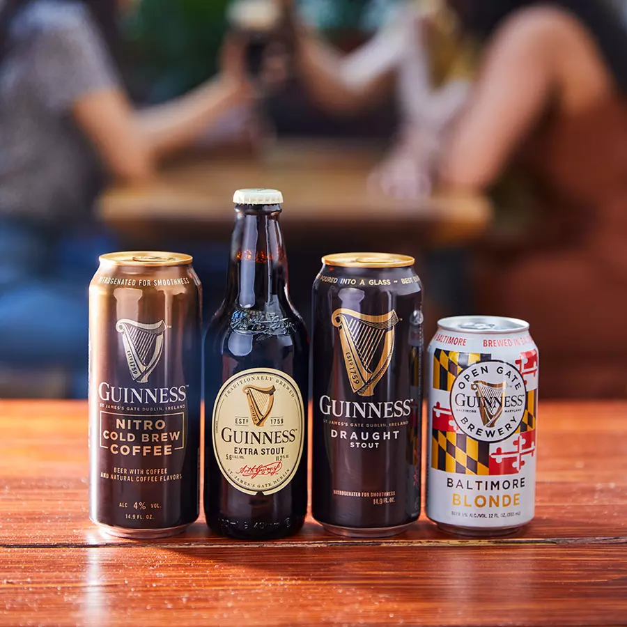 Four Guinness beer varieties—Nitro Cold Brew Coffee, Extra Stout, Draught Stout, and Baltimore Blonde—are displayed on a wooden table with blurred people in the background.
