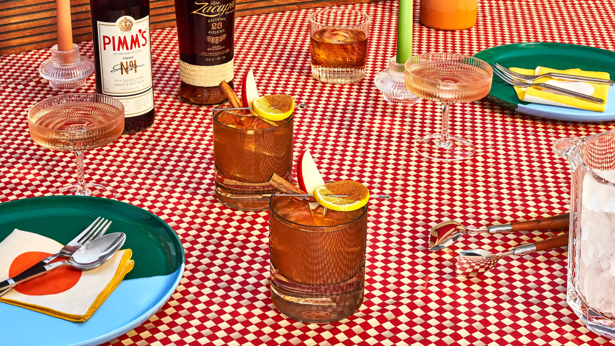 Several cocktails with fruit garnishes, bottles of liquor, plates with utensils, and napkins are arranged on a red and white checkered tablecloth.