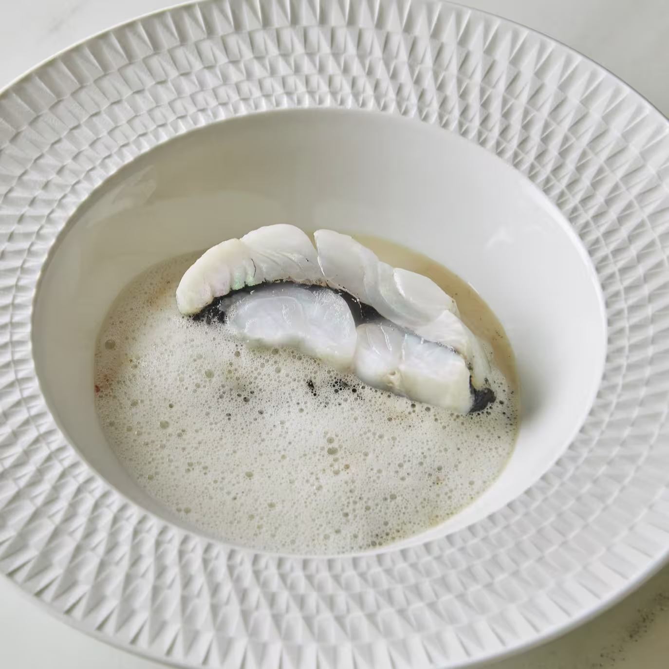Plato sofisticado con pescado blanco delicadamente presentado sobre una espuma ligera, servido en un elegante cuenco de diseño geométrico.