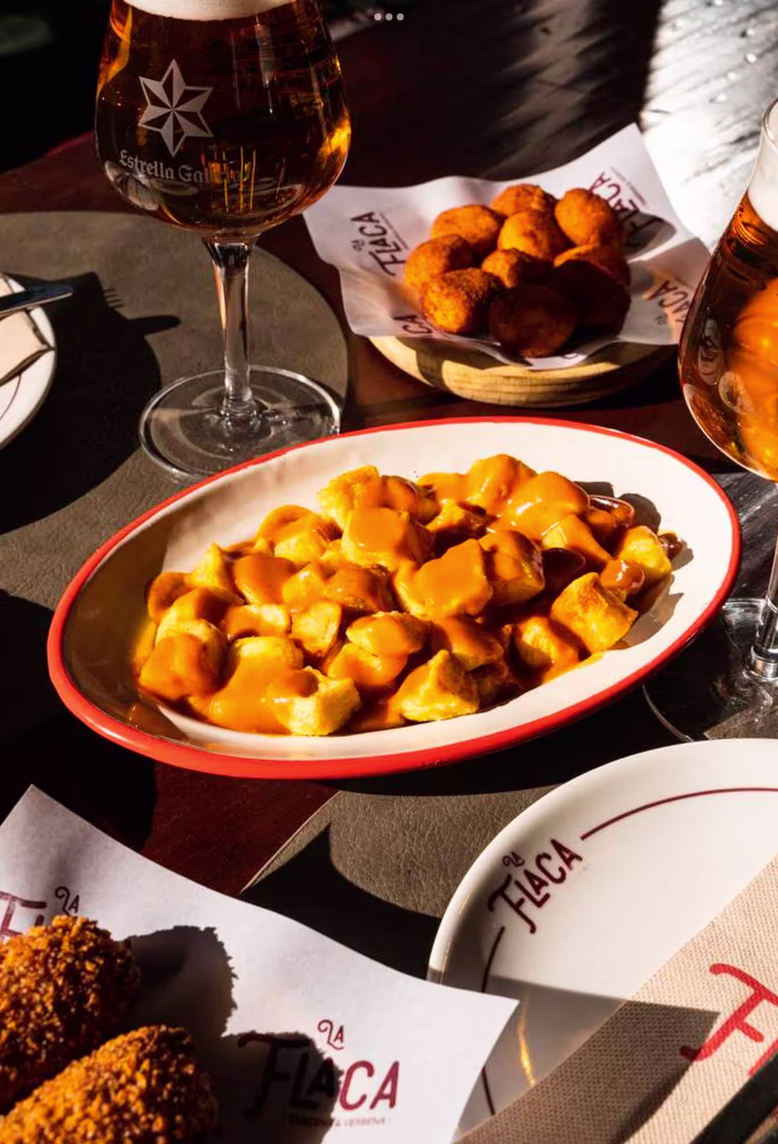 La foto muestra una mesa en La flaca con varios platos y un vaso de cerveza. En primer plano, hay un plato de patatas bravas, un plato tradicional español de tapas que consiste en papas fritas con una salsa de tomate picante.