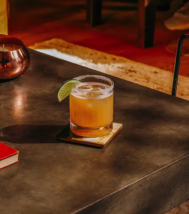 A cocktail garnished with a lime wedge sits on a square coaster on a dark table, with a red book and a candle nearby.