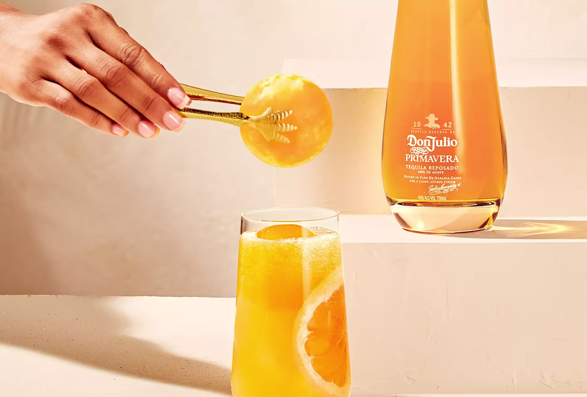 A hand uses tongs to place an orange slice into a glass of orange drink beside a Don Julio Primavera tequila bottle.