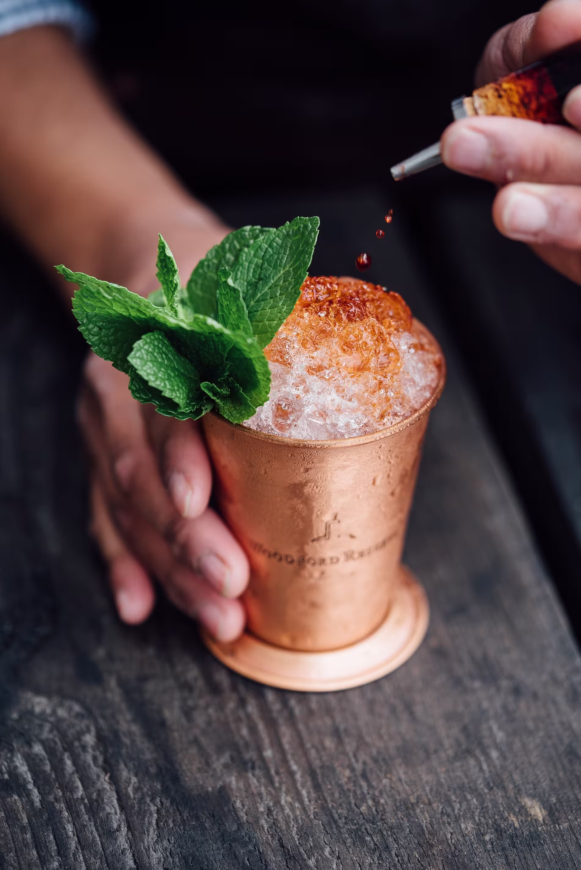 A hand holds a copper cup filled with crushed ice, garnished with fresh mint, while another hand adds drops of bitters to the drink.