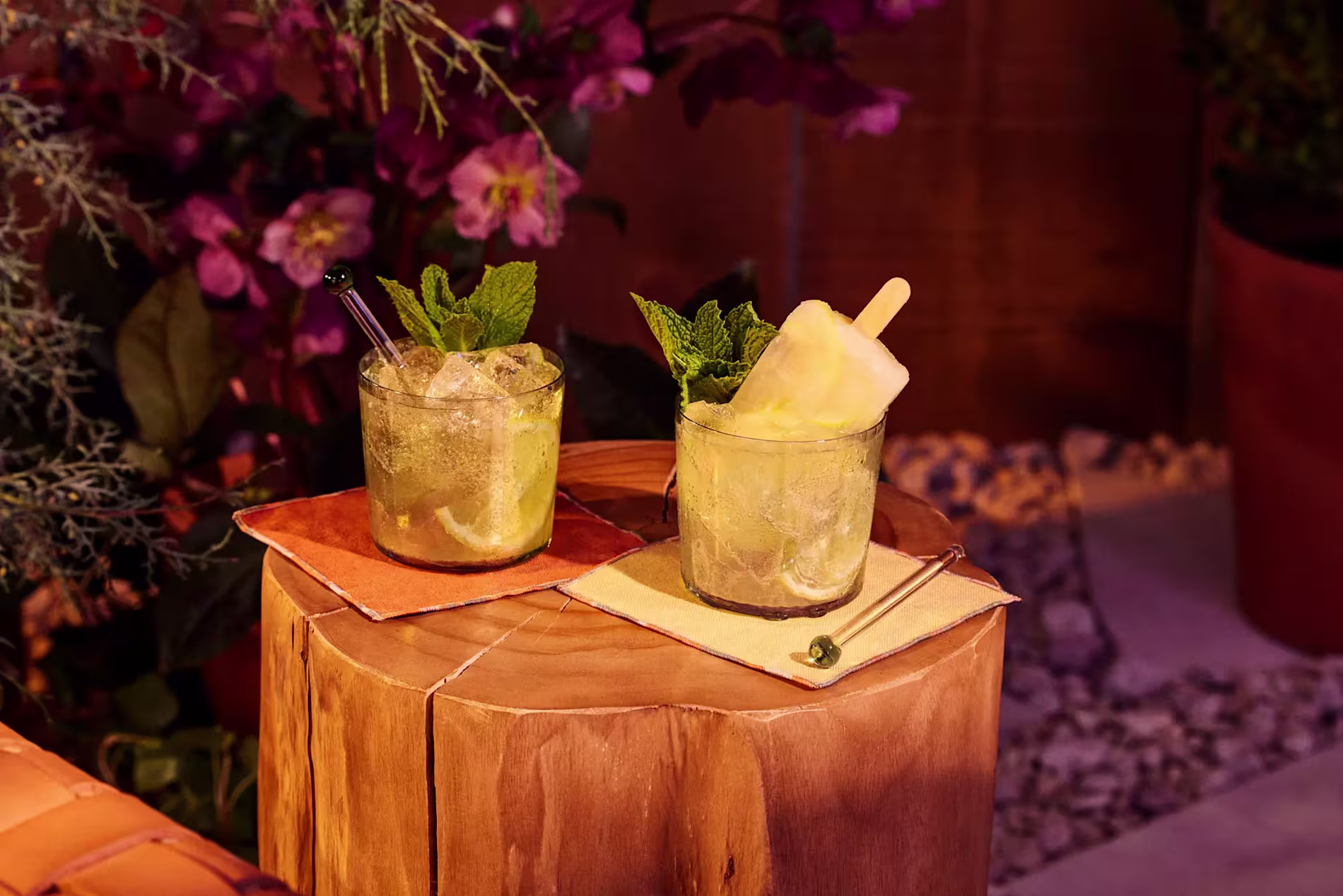 Two cocktails with lime and mint, one with a popsicle, are placed on a wooden stump with napkins, surrounded by flowers and plants.