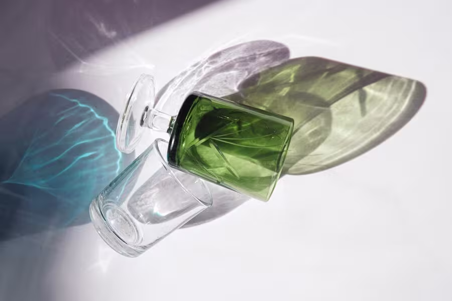 A clear glass and a green glass cup are lying on their sides on a white surface, casting blue and green shadows.