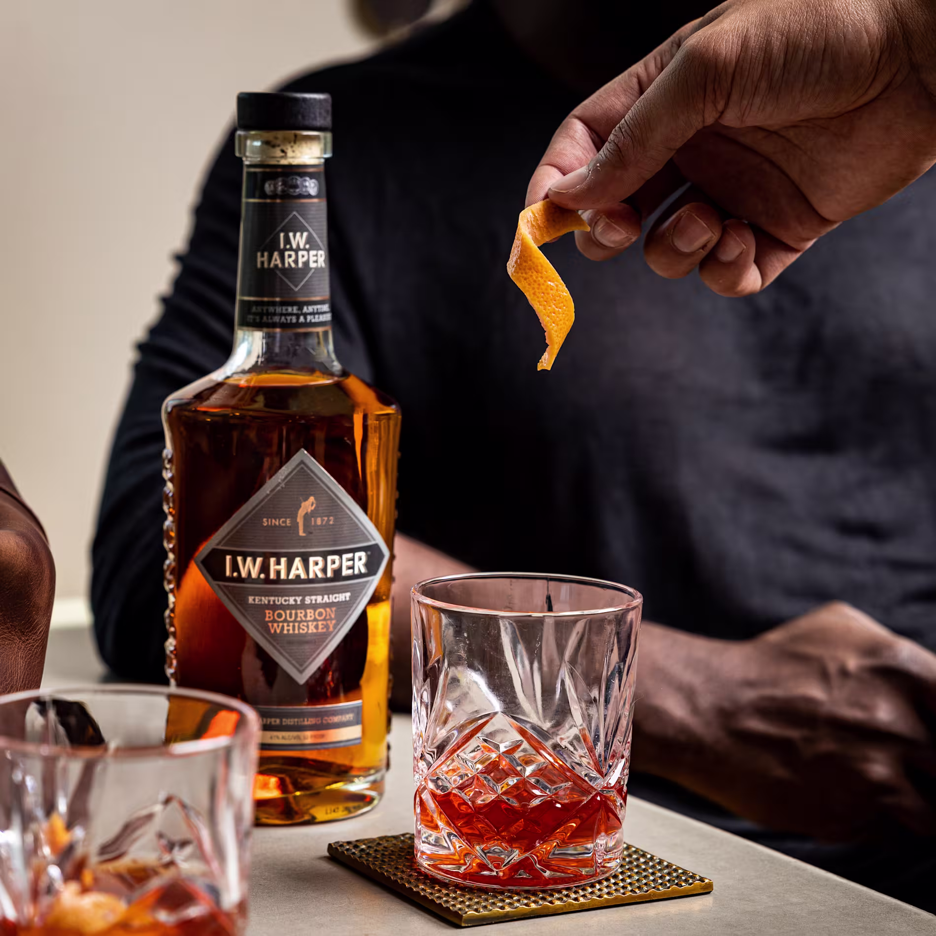 A hand garnishes a glass of bourbon cocktail with an orange twist next to a bottle of I.W. Harper bourbon whiskey on a table.