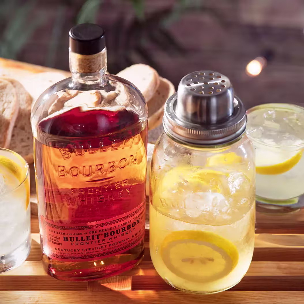A bottle of Bulleit Bourbon whiskey next to a mason jar cocktail shaker with lemon slices, surrounded by cocktails and bread on a wooden surface.
