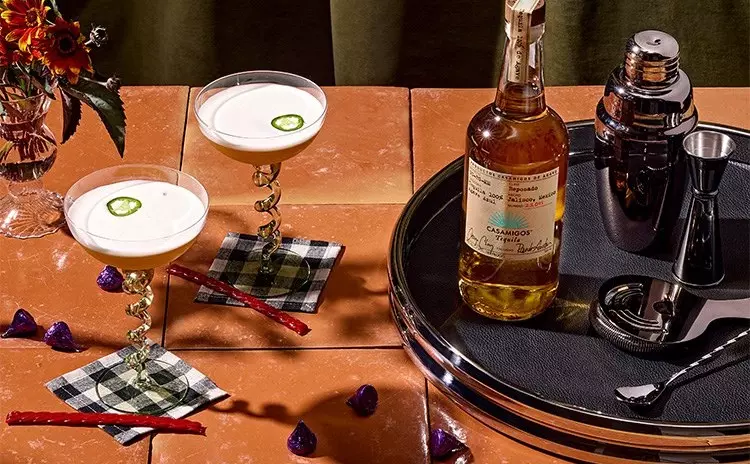 Two cocktails with frothy tops and jalapeño slices, a bottle of tequila, cocktail shaker, and bar tools arranged on a tray atop a tiled table with flowers and candy nearby.