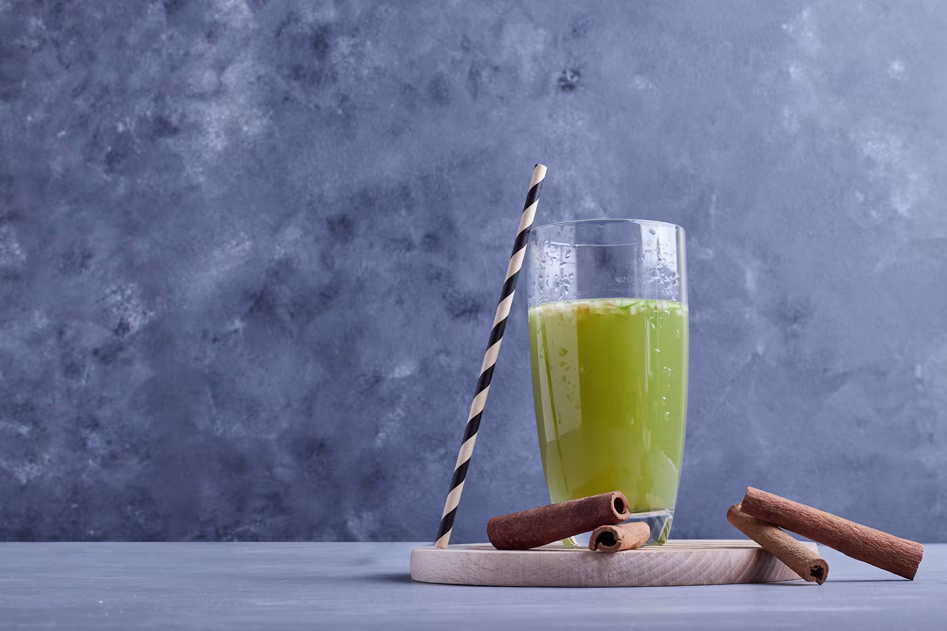 A glass of green juice with a striped straw sits on a round wooden tray next to several cinnamon sticks, against a gray textured background.
