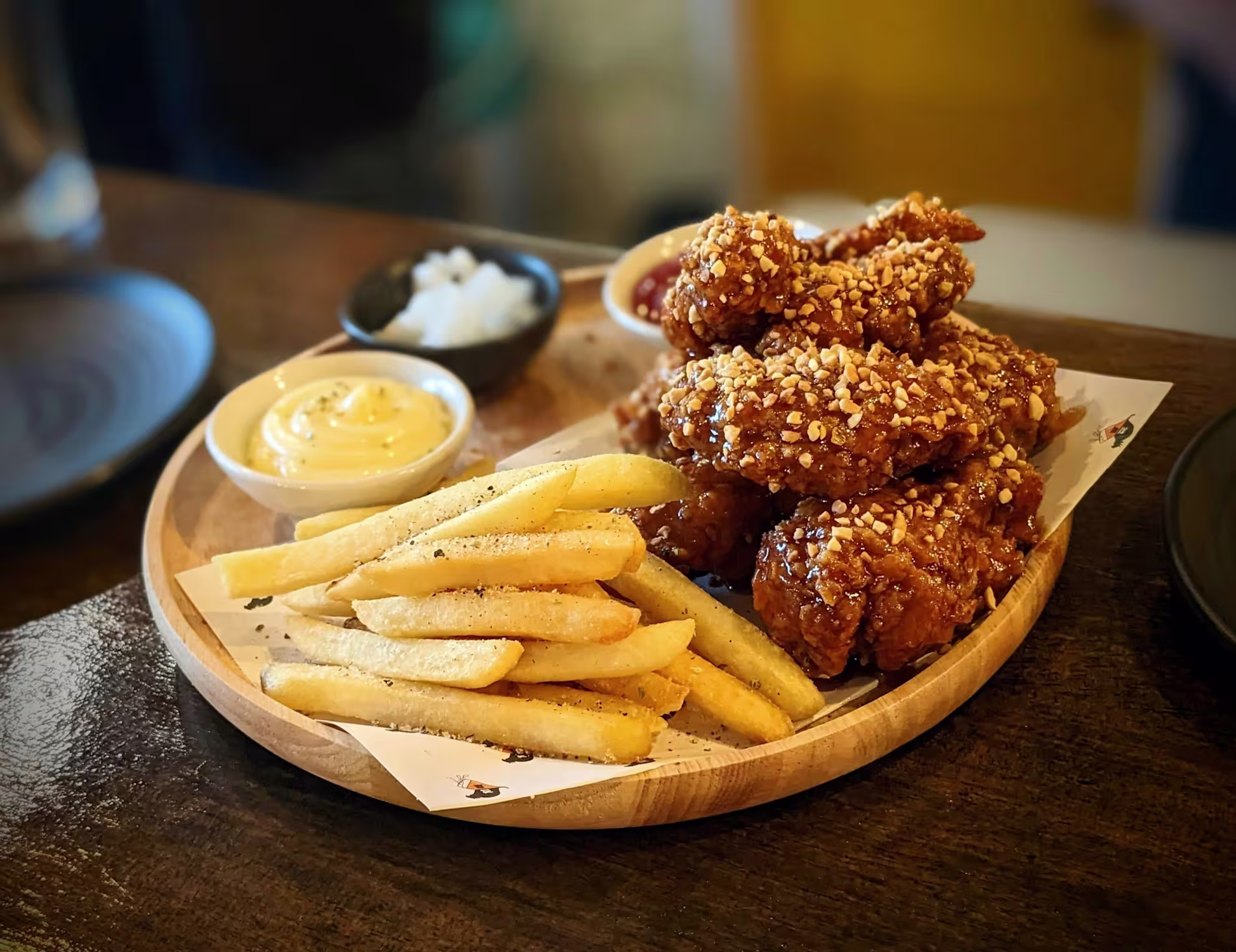 A wooden plate with seasoned French fries, crispy fried chicken wings topped with sesame seeds, a dish of mayonnaise, and a small serving of pickled radish.