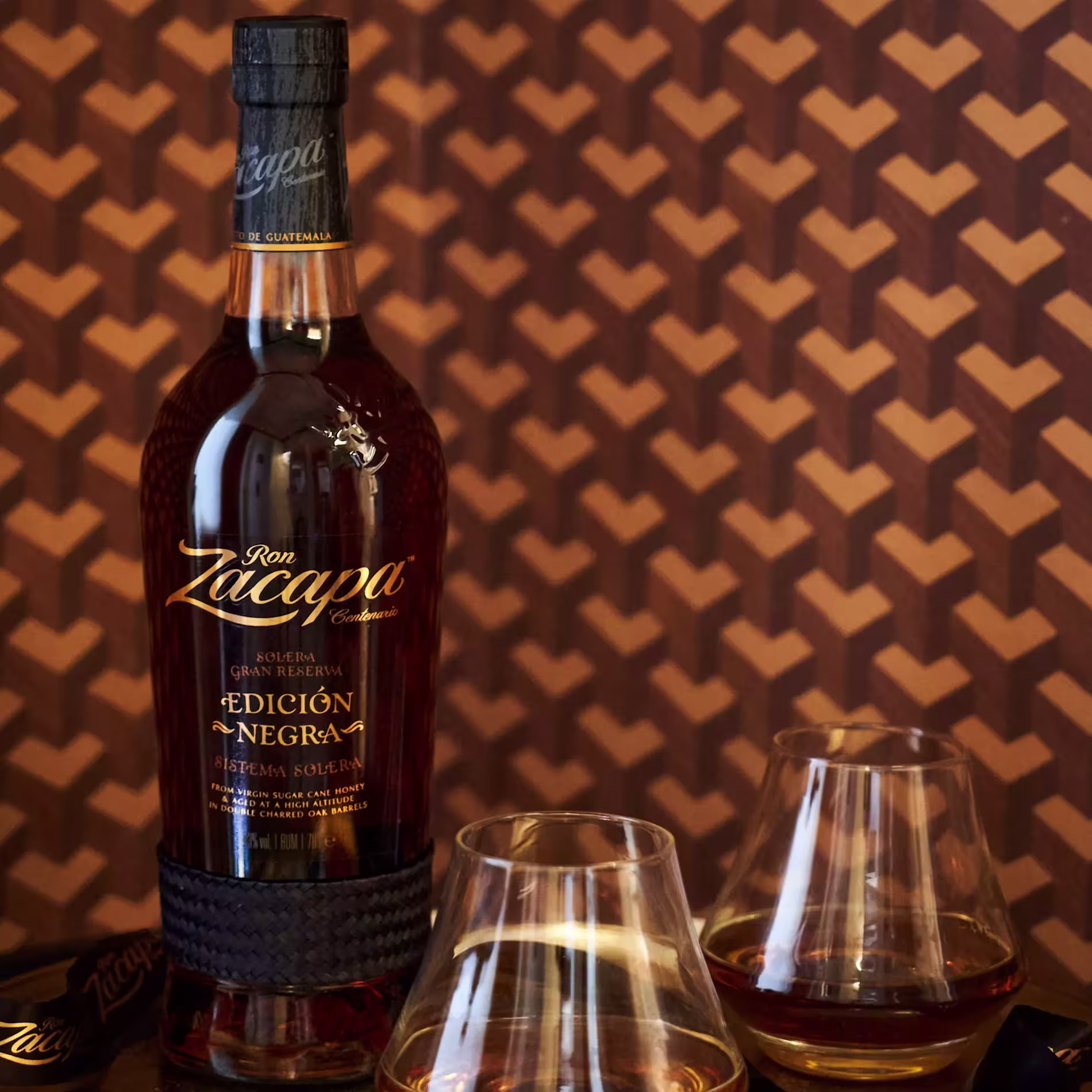 A bottle of Ron Zacapa Edición Negra rum stands next to two filled glasses on a table with a geometric brown patterned background.
