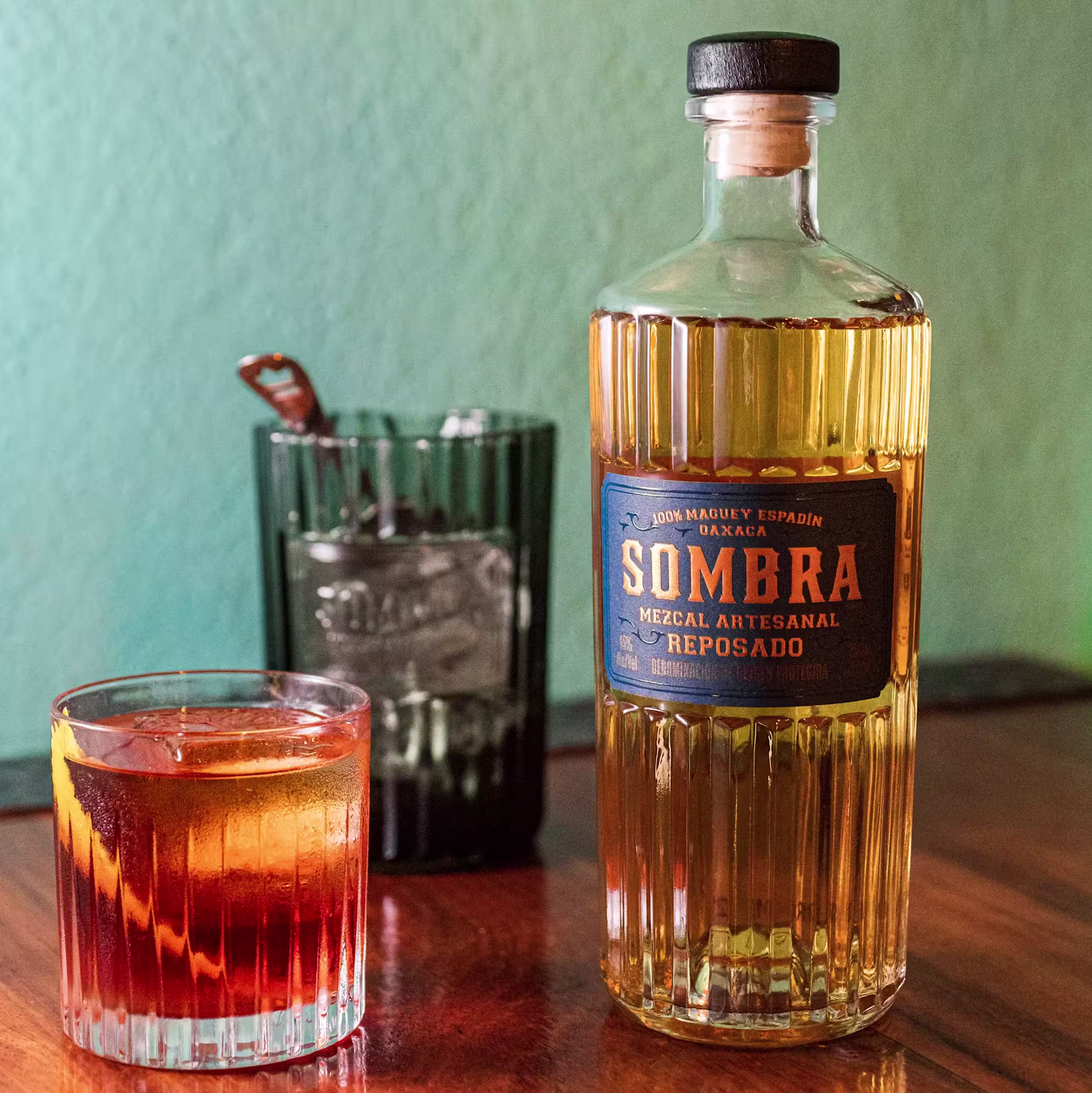 A bottle of Sombra Mezcal Reposado sits on a wooden table next to a glass of amber cocktail and a green glass with a metal spoon inside.