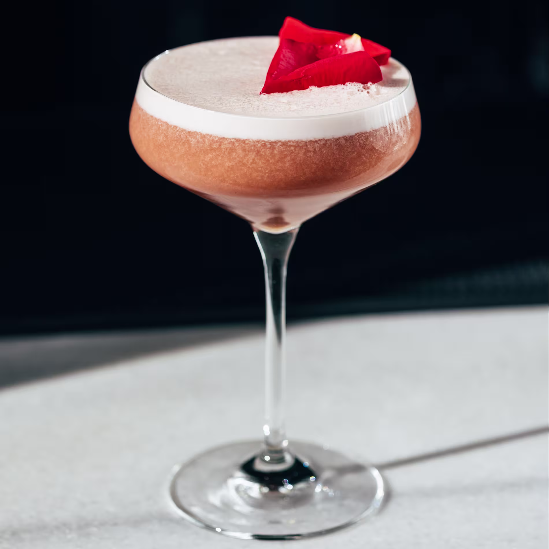 La foto muestra un cóctel en un vaso con tallo, un vaso de martini o coupe. El cóctel tiene una capa espumosa en la parte superior y está adornado con una pieza de color rojo brillante.