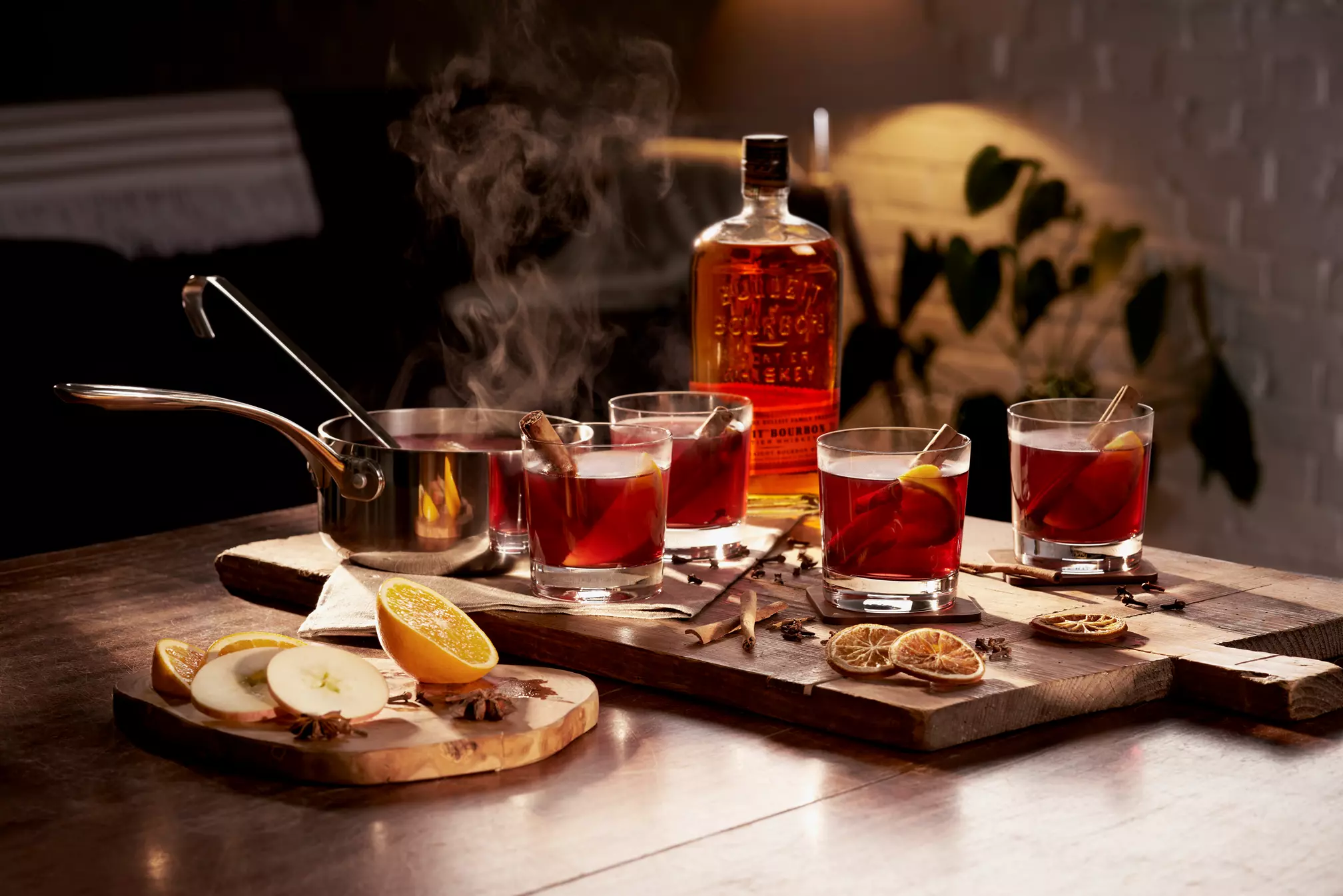 A bottle of whiskey, three glasses of mulled cocktail with cinnamon sticks, a steaming saucepan, and sliced fruits are arranged on a wooden table.