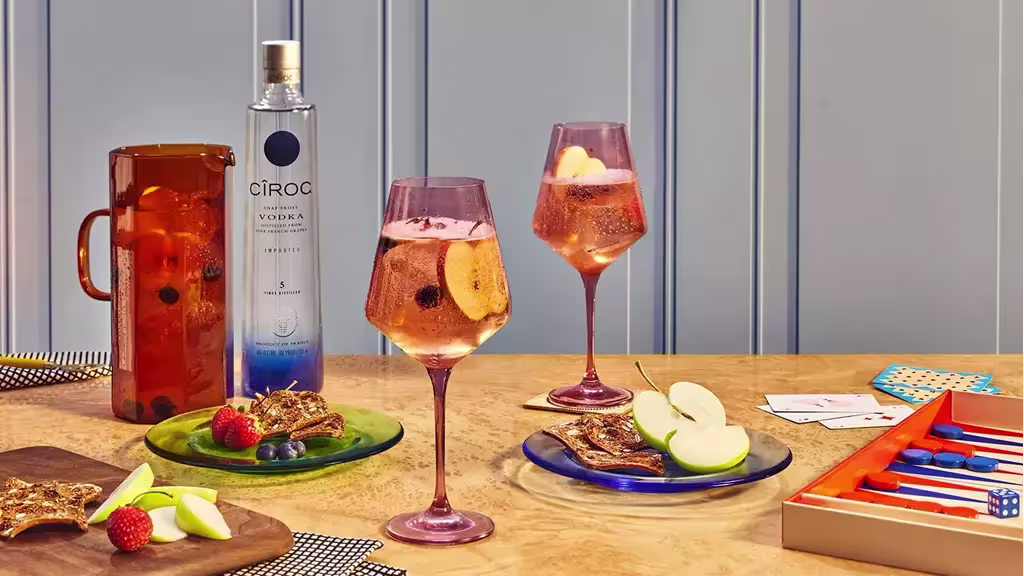 A table with fruit, snacks, two glasses of pink cocktail, a bottle of Ciroc vodka, a pitcher, and a backgammon game set.