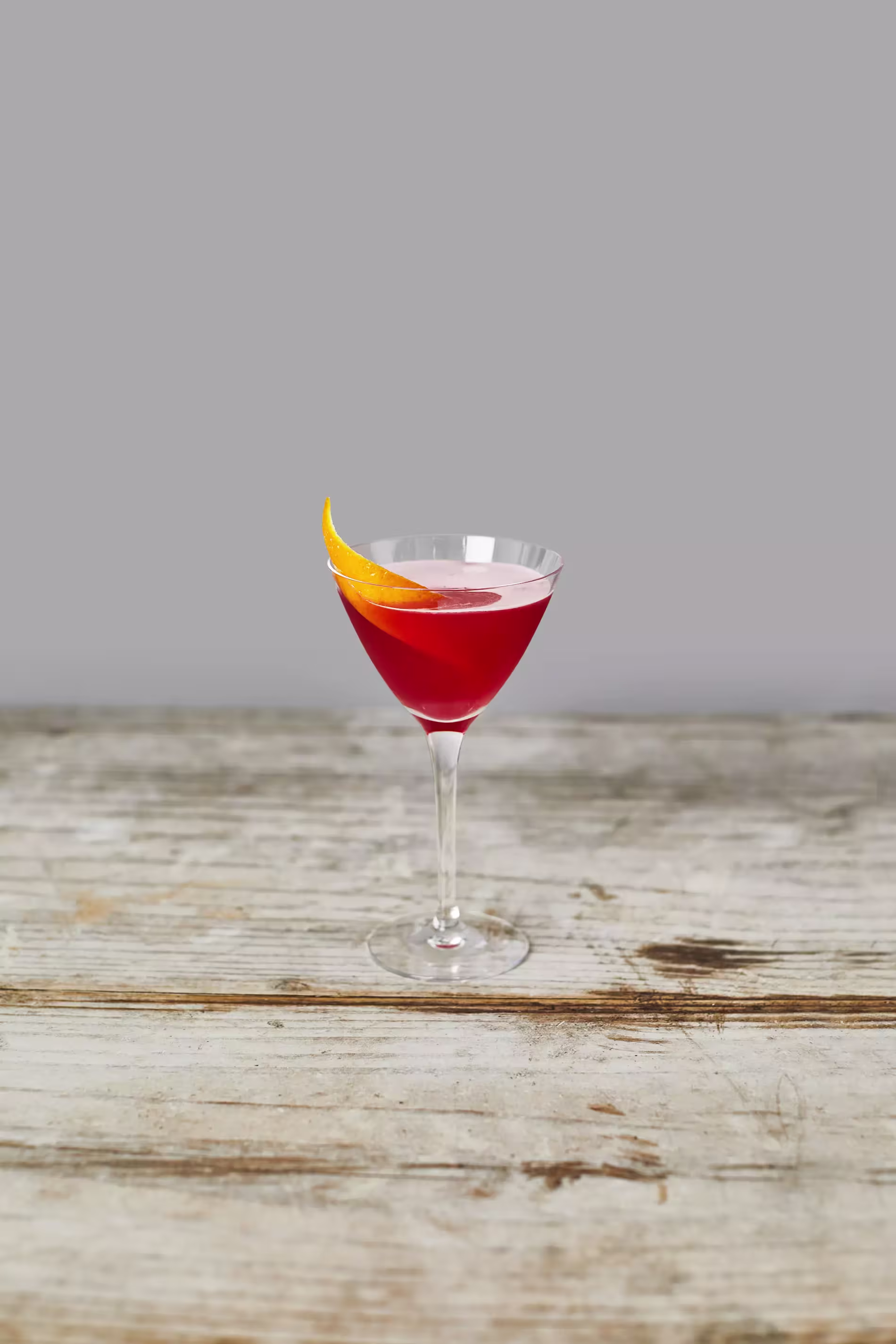A stemmed glass filled with a red cocktail, garnished with a twisted orange peel, placed on a rustic wooden surface against a plain gray background.
