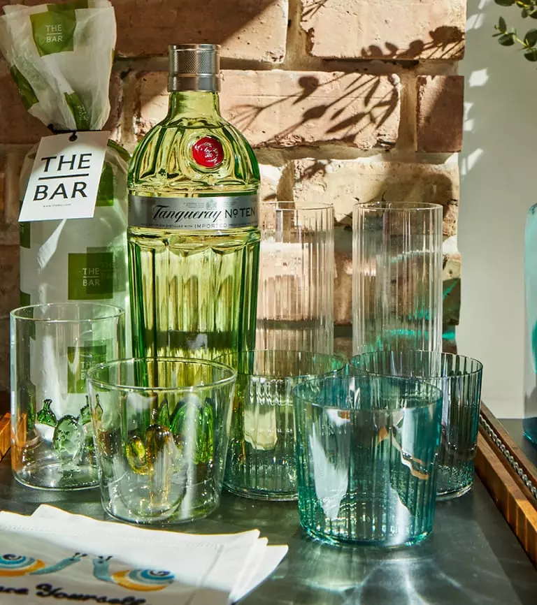 Tanqueray No. Ten gin bottle surrounded by various empty textured glasses on a table, with a brick wall and wrapped package in the background.