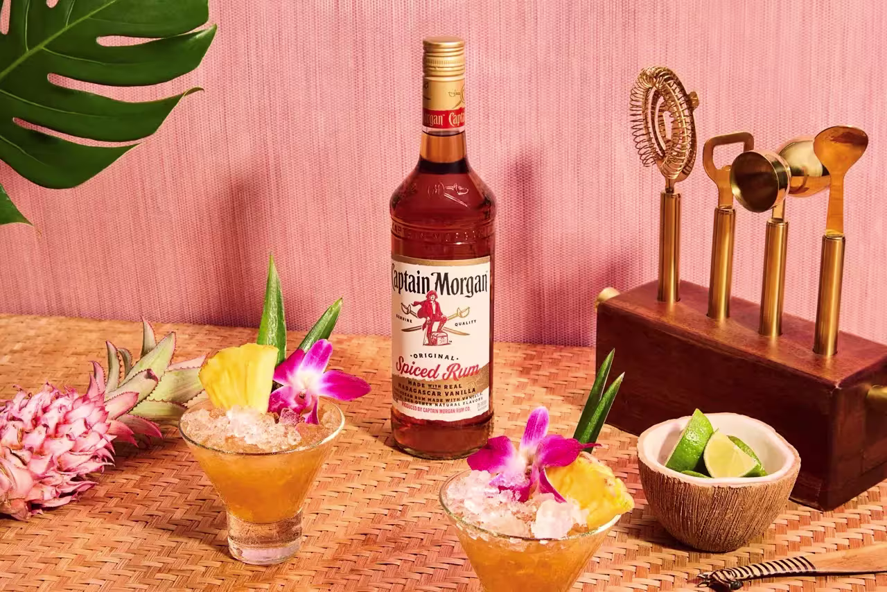 A bottle of Captain Morgan spiced rum with two tropical cocktails, garnished with pineapple and flowers, sits on a woven mat beside bar tools, a coconut bowl of lime wedges, and a pink pineapple.