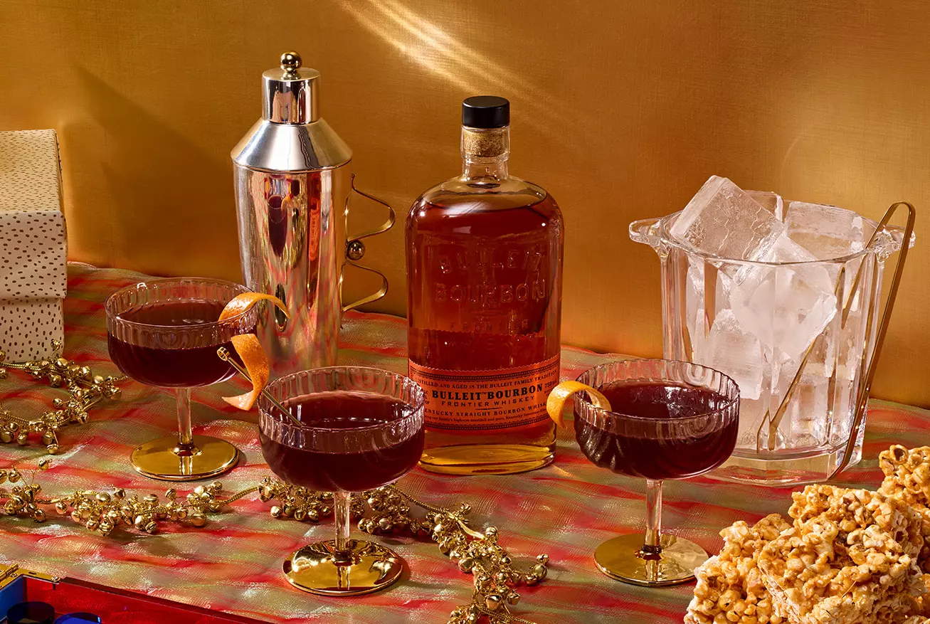 A bottle of Bulleit Bourbon, three cocktails with orange twists, a cocktail shaker, an ice bucket, and caramel popcorn on a festive tablecloth with gift boxes in the background.
