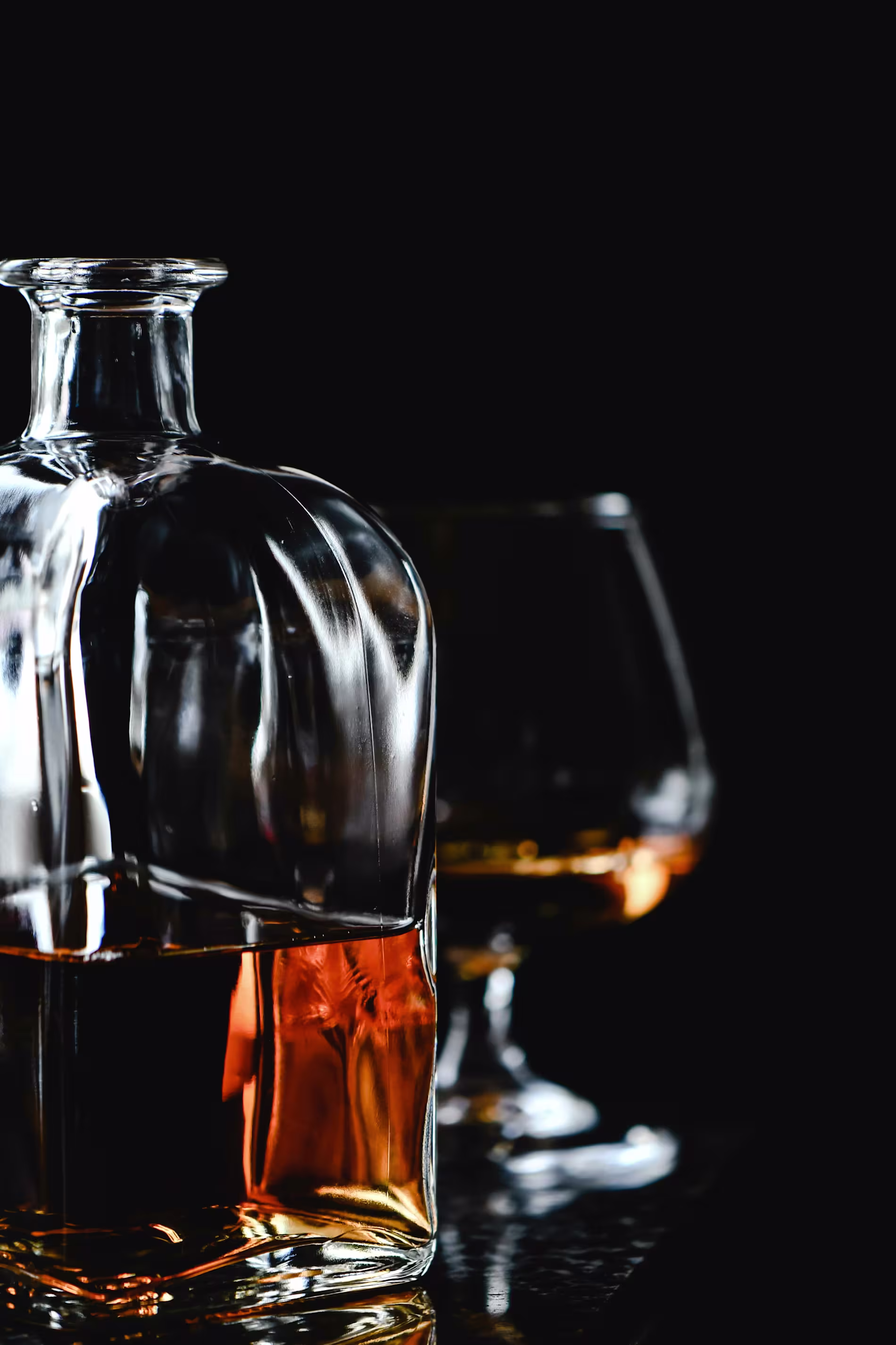 A glass decanter filled with amber-colored liquor sits next to a filled snifter glass against a dark background.