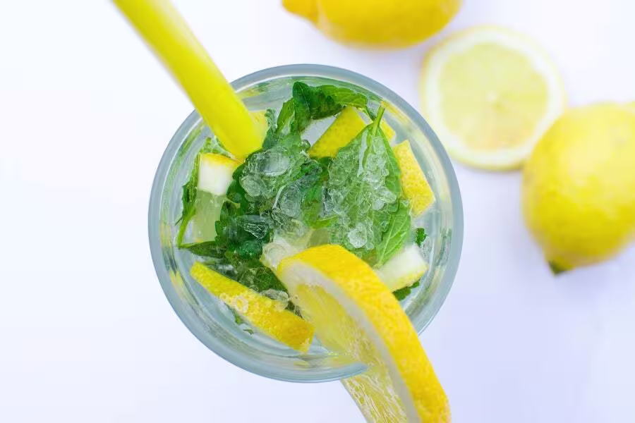 Top view of a glass of lemonade with ice, lemon slices, mint leaves, and a yellow straw, with whole and sliced lemons in the background.