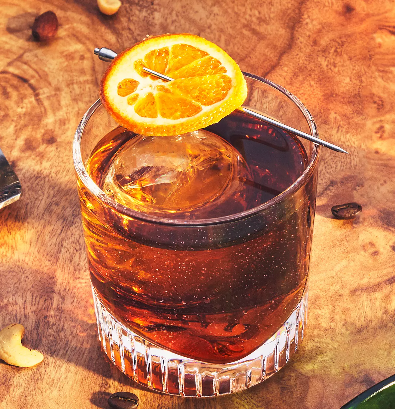 A glass of amber-colored cocktail with a large ice sphere, garnished with a slice of orange on a metal pick, sits on a wooden surface.