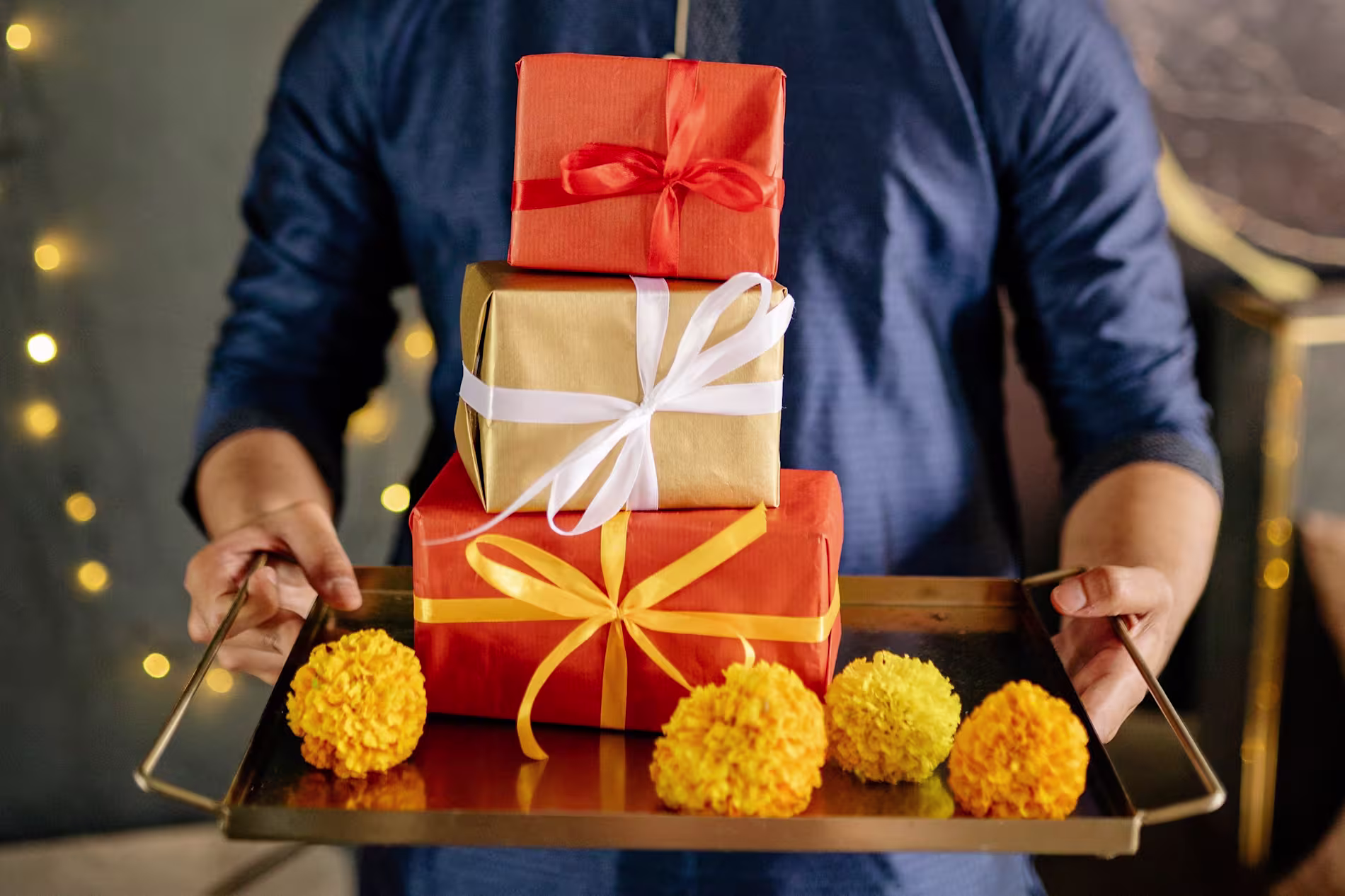 A person in a blue outfit holds a tray with three wrapped gift boxes in orange and gold, tied with ribbons, and decorated with yellow marigold flowers.
