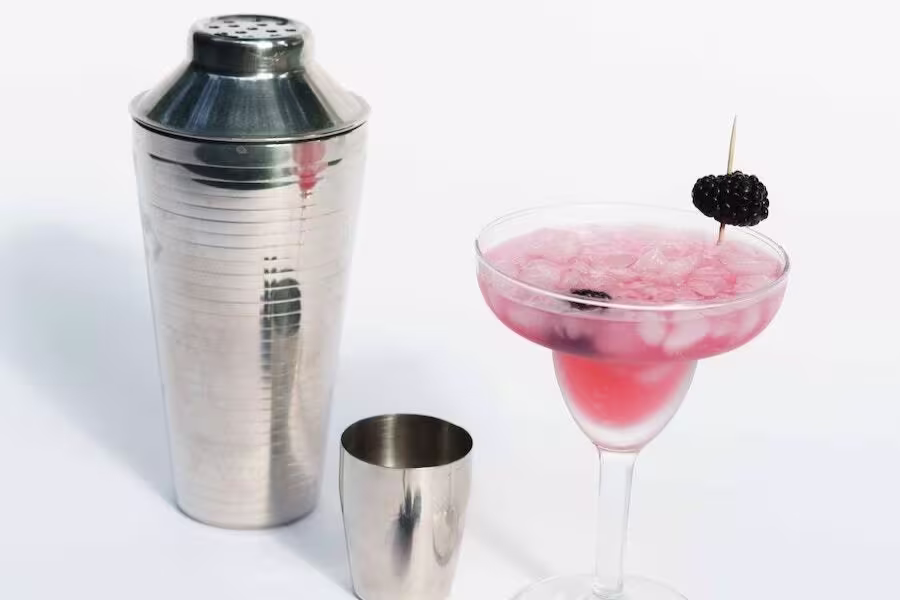 A metal cocktail shaker and jigger are placed next to a pink cocktail in a stemmed glass, garnished with a blackberry on a toothpick.