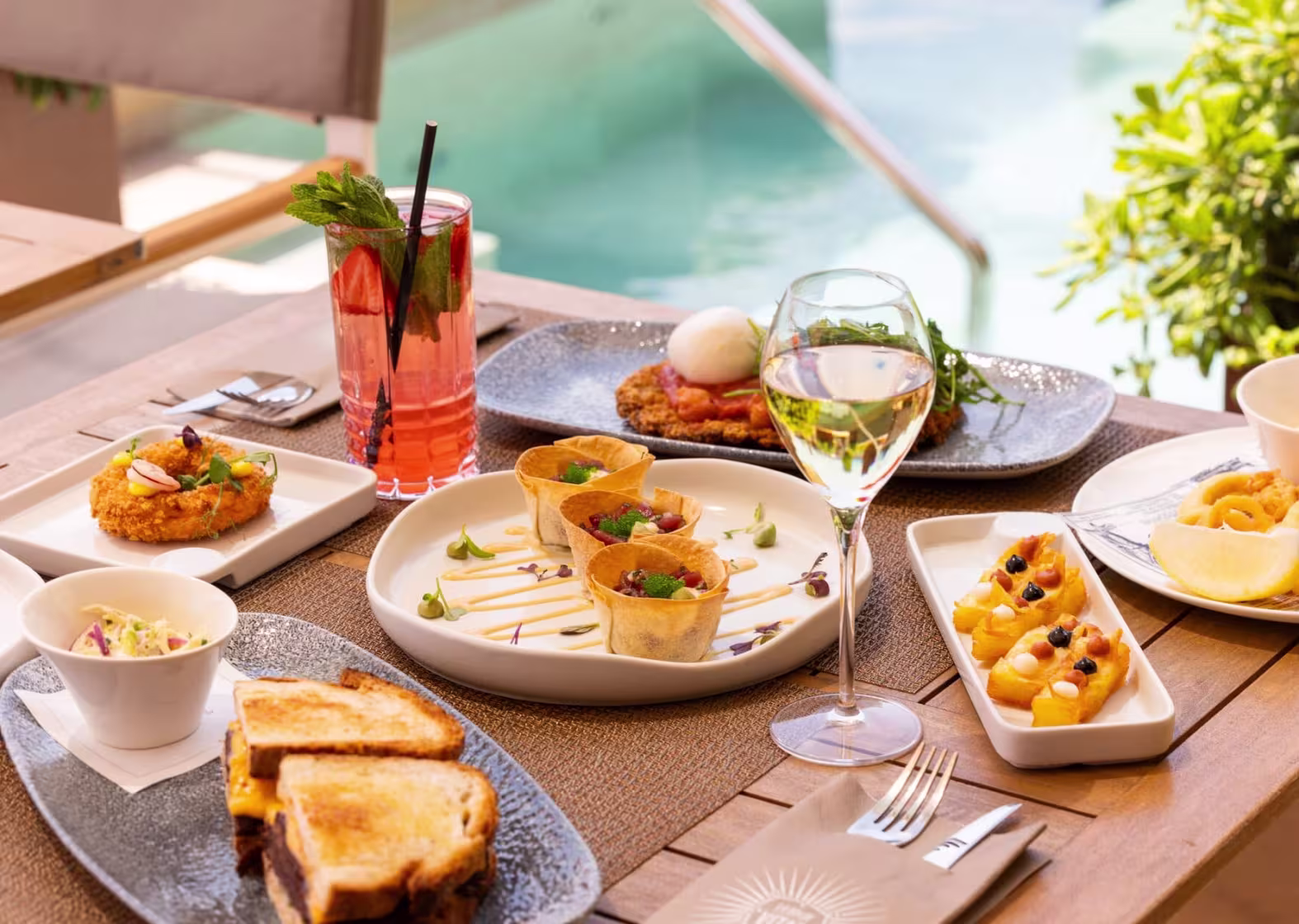 Mesa al aire libre en La Dolce Vitae con variedad de platos gourmet, cócteles y vino, junto a una piscina y vegetación fresca.