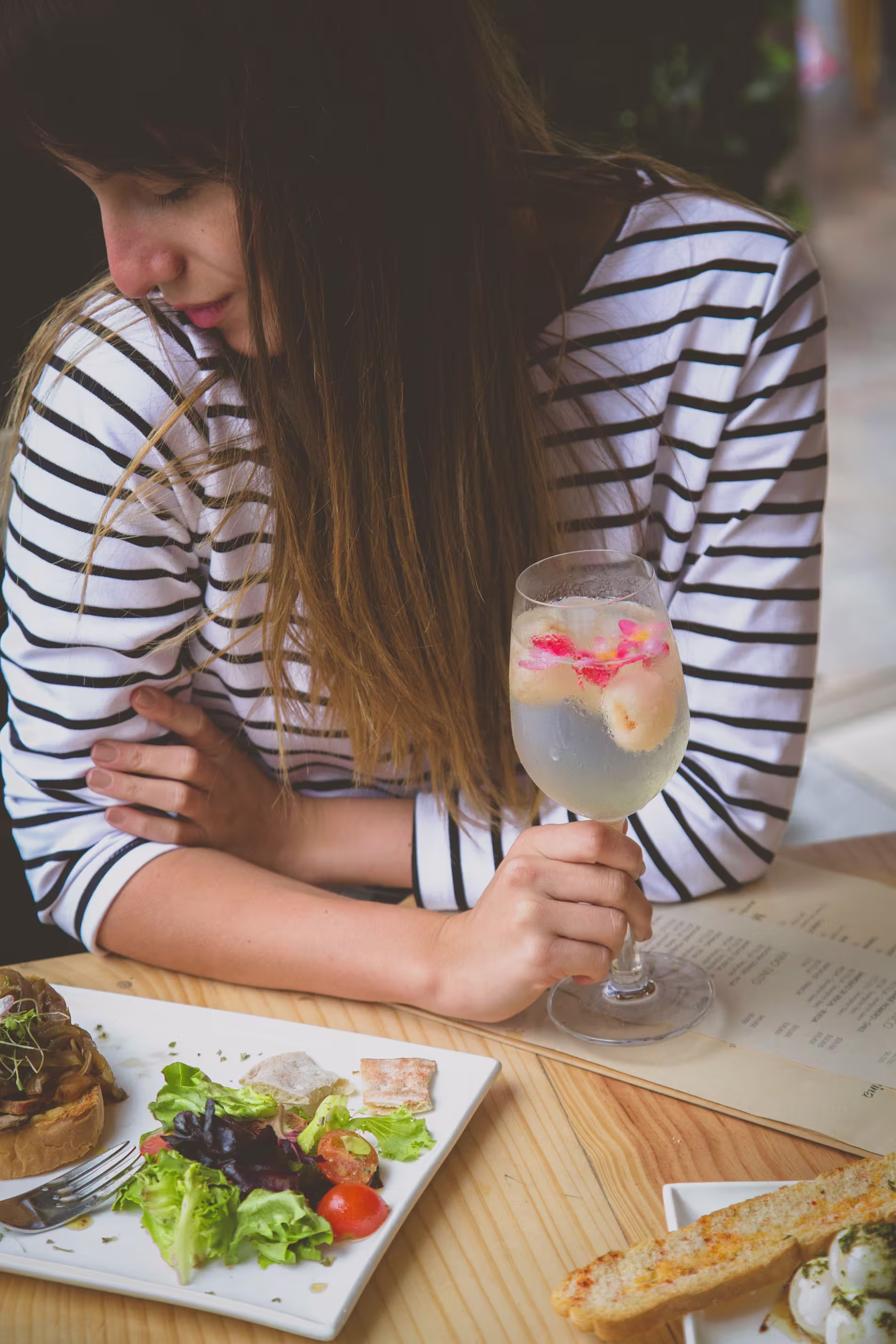 A woman in a striped shirt holds a glass of clear beverage with fruit and flowers, seated at a table with salad, bread, and other food.