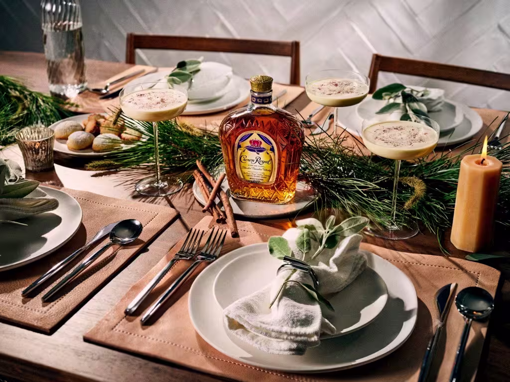A holiday table set with plates, cutlery, four glasses of eggnog, a bottle of Crown Royal whisky, greenery, candles, and festive napkins.