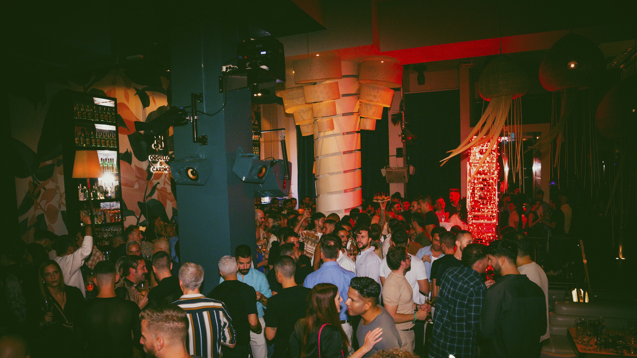 La imagen muestra el interior de un bar animado y lleno de gente disfrutando de la noche. Con una iluminación cálida en tonos rojos y una decoración original, el ambiente invita a socializar en un espacio vibrante y moderno.