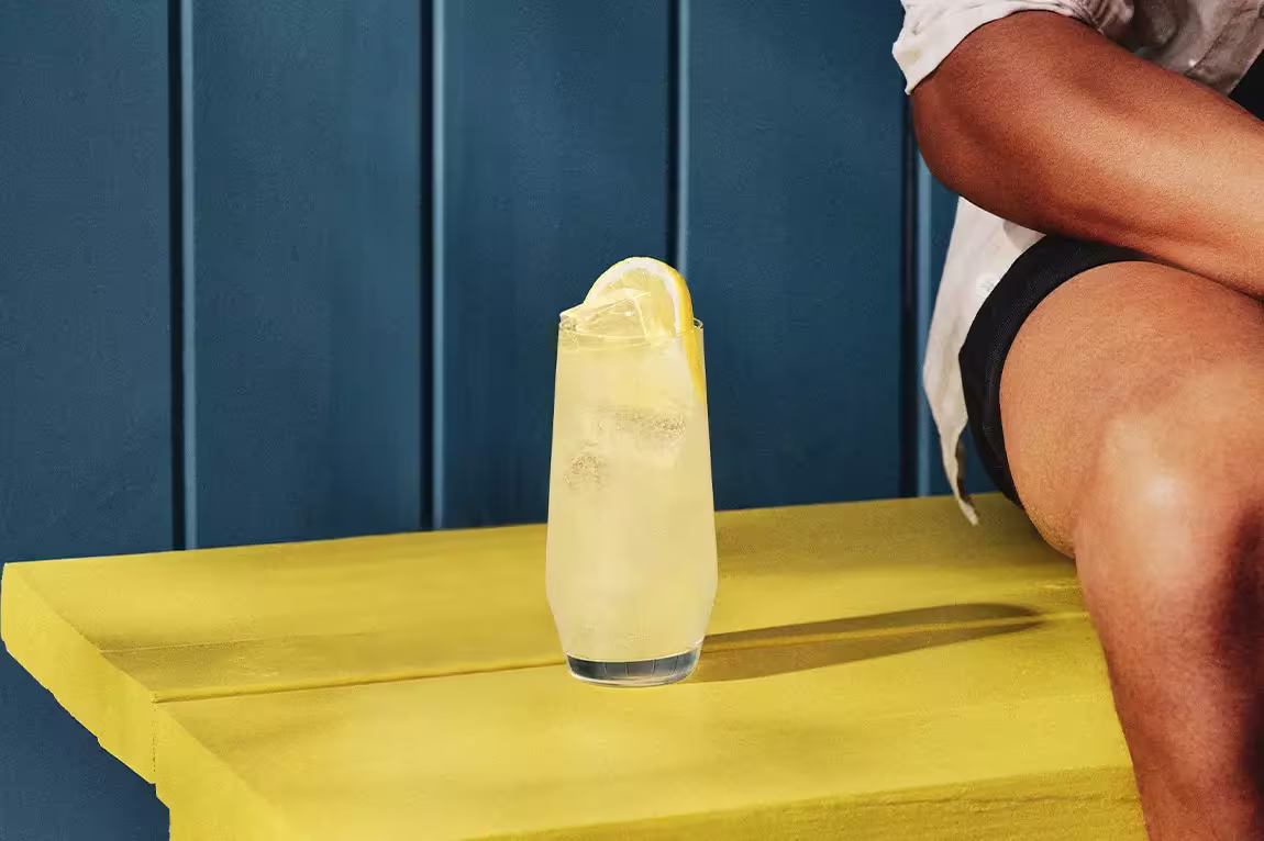 A glass of iced lemonade with a lemon slice sits on a yellow table next to a person wearing a white shirt and black shorts.
