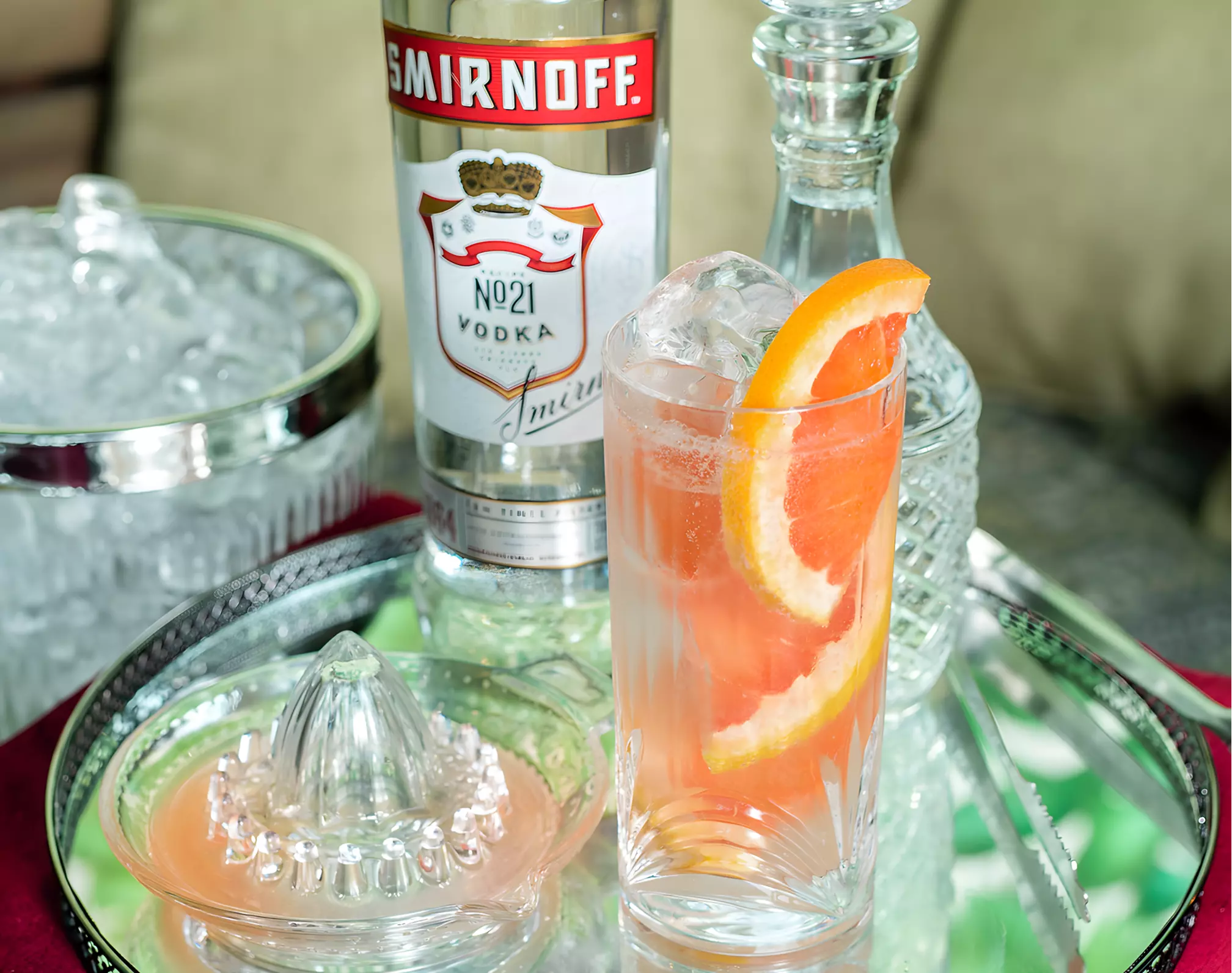 A glass of grapefruit cocktail with ice and orange slices sits on a tray with a bottle of Smirnoff vodka, a decanter, a juicer, and a bowl of ice.