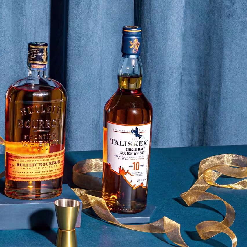 Two bottles of whiskey, Bulleit Bourbon and Talisker 10-Year-Old Single Malt, stand on a blue surface with a gold ribbon and jigger, against a blue curtain backdrop.