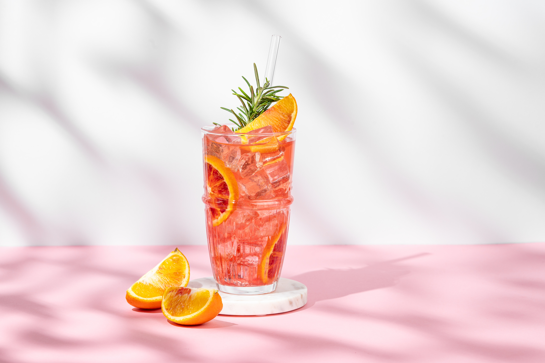A glass of pink citrus drink with ice, garnished with orange slices and rosemary, sits on a white coaster. Two orange wedges rest on the pink surface beside the glass.