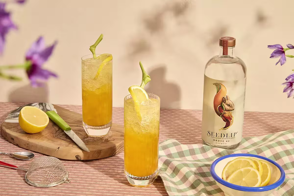 Two tall glasses with orange cocktails and lemon slices sit on a checkered cloth next to a Seedlip Grove 42 bottle, a bowl of lemon slices, a strainer, and a halved lemon on a cutting board.