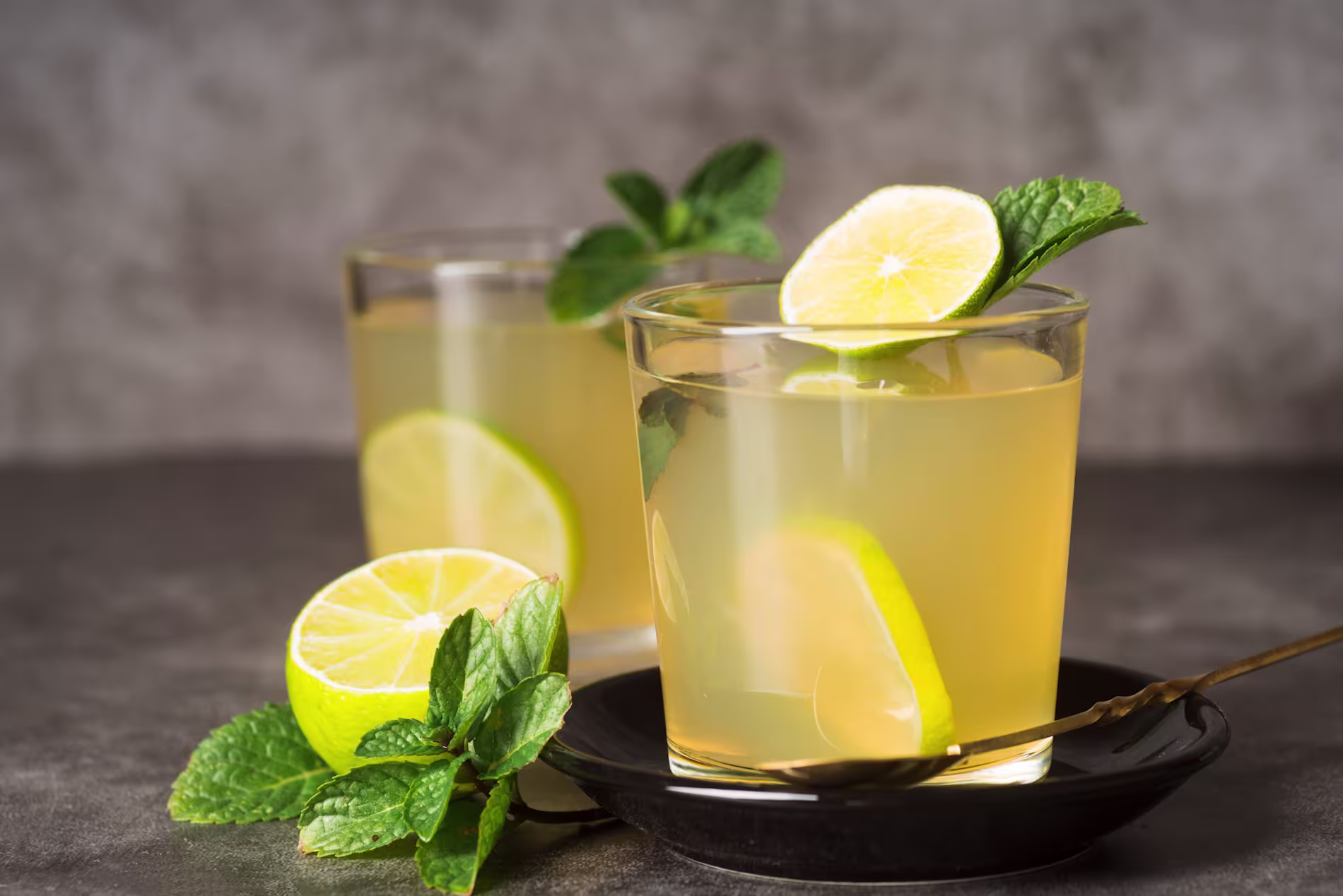 Two glasses of lime and mint beverage with lime slices and mint sprigs, one glass on a black plate with a spoon, set on a dark surface.