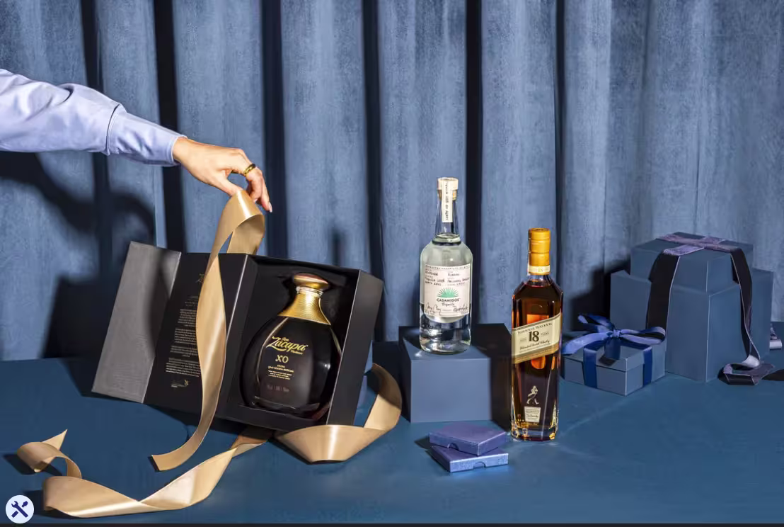 Una persona preparando paquetes de regalo de tres botellas, sobre fondo azul, con varias cajas de regalo y lazo dorado.