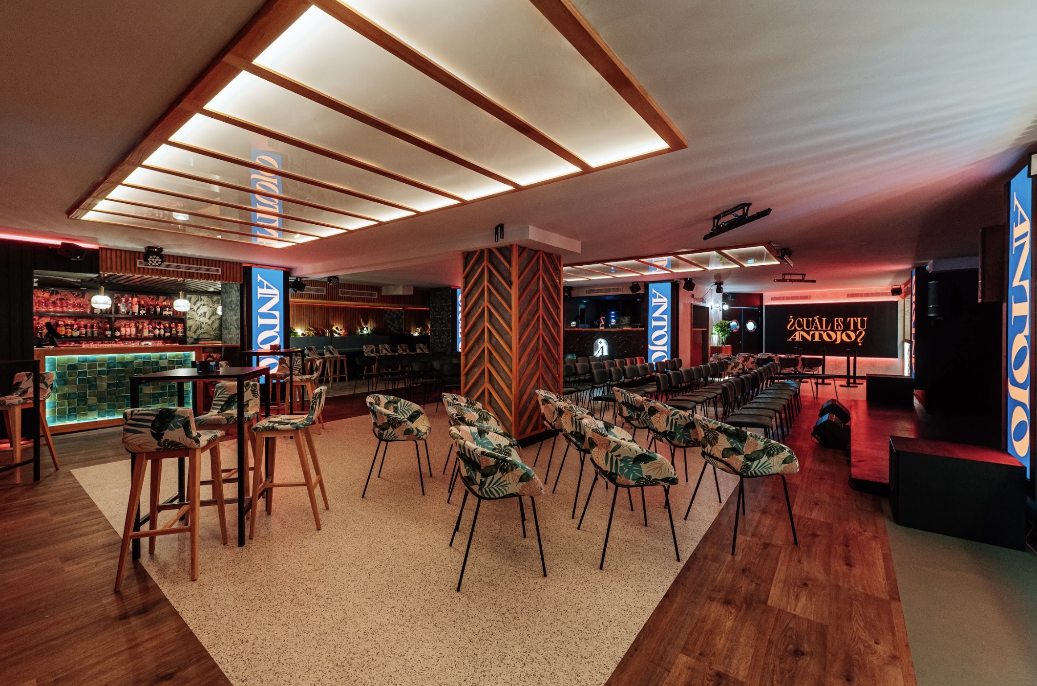 Interior de Antojo, con un diseño moderno que combina tonos cálidos, un bar iluminado y asientos con estampados tropicales.