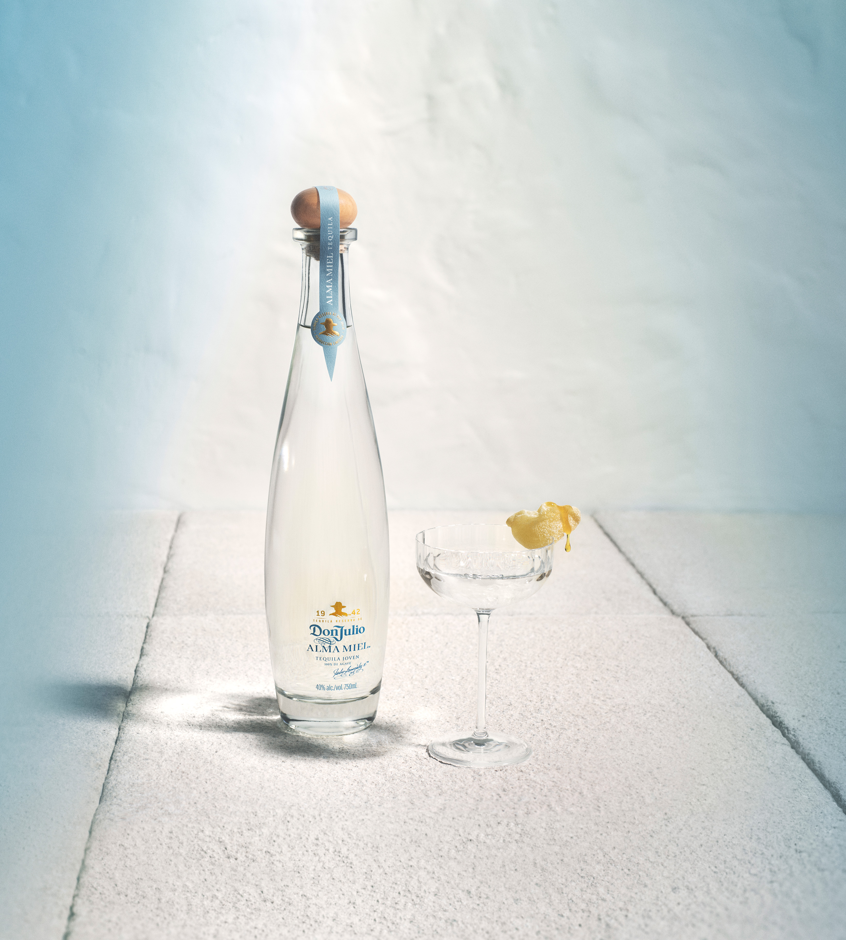 A clear glass bottle of water with a cork top stands next to a cocktail glass garnished with a lemon twist on a white textured surface.