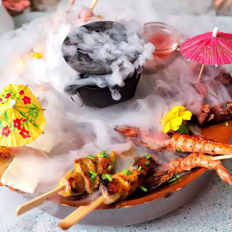 Bandeja con brochetas, flores, sombrillas y olla humeante en el centro, rodeada de niebla de hielo seco.