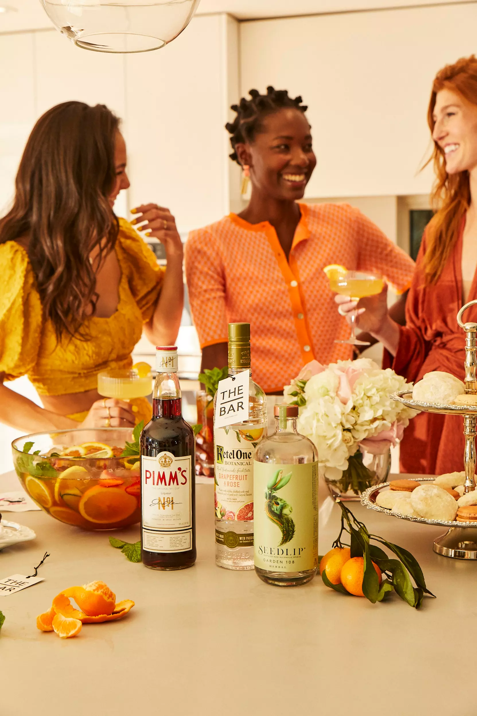 Three women stand around a kitchen island with cocktails, bottles of beverages, a punch bowl, flowers, and pastries arranged on the countertop.