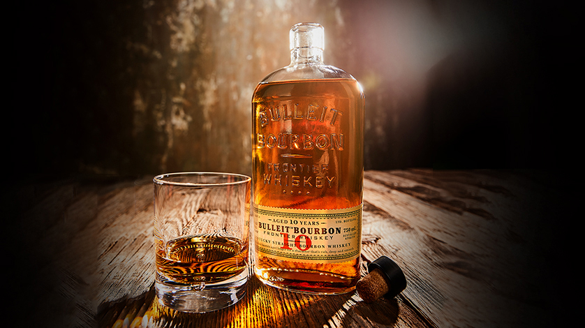 A bottle of Bulleit Bourbon whiskey stands next to a glass of poured whiskey and a bottle cap on a wooden surface, with warm lighting in the background.