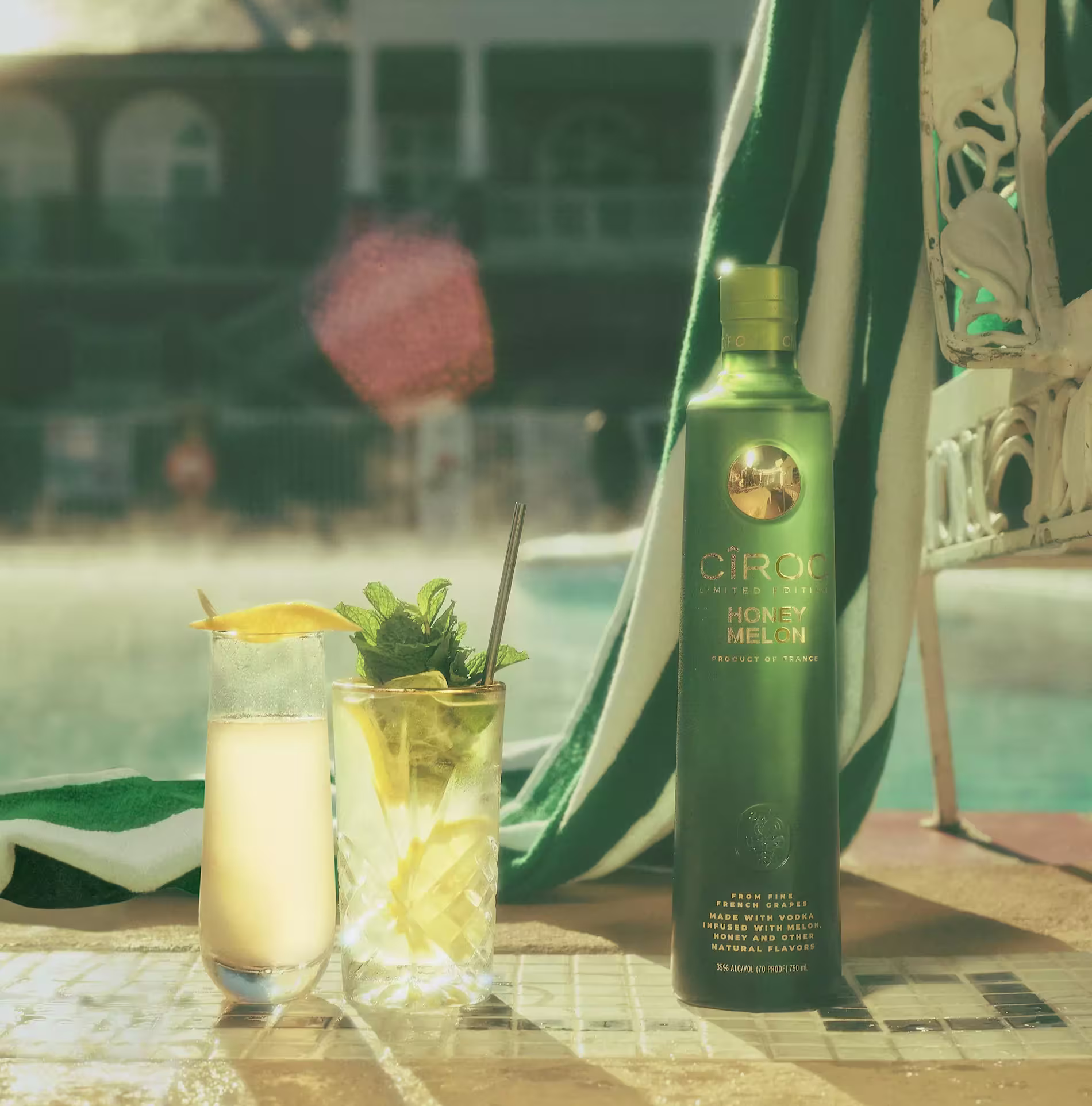 A bottle of Ciroc Honey Melon vodka stands beside two cocktails with garnishes by a poolside with sunlight and a towel in the background.