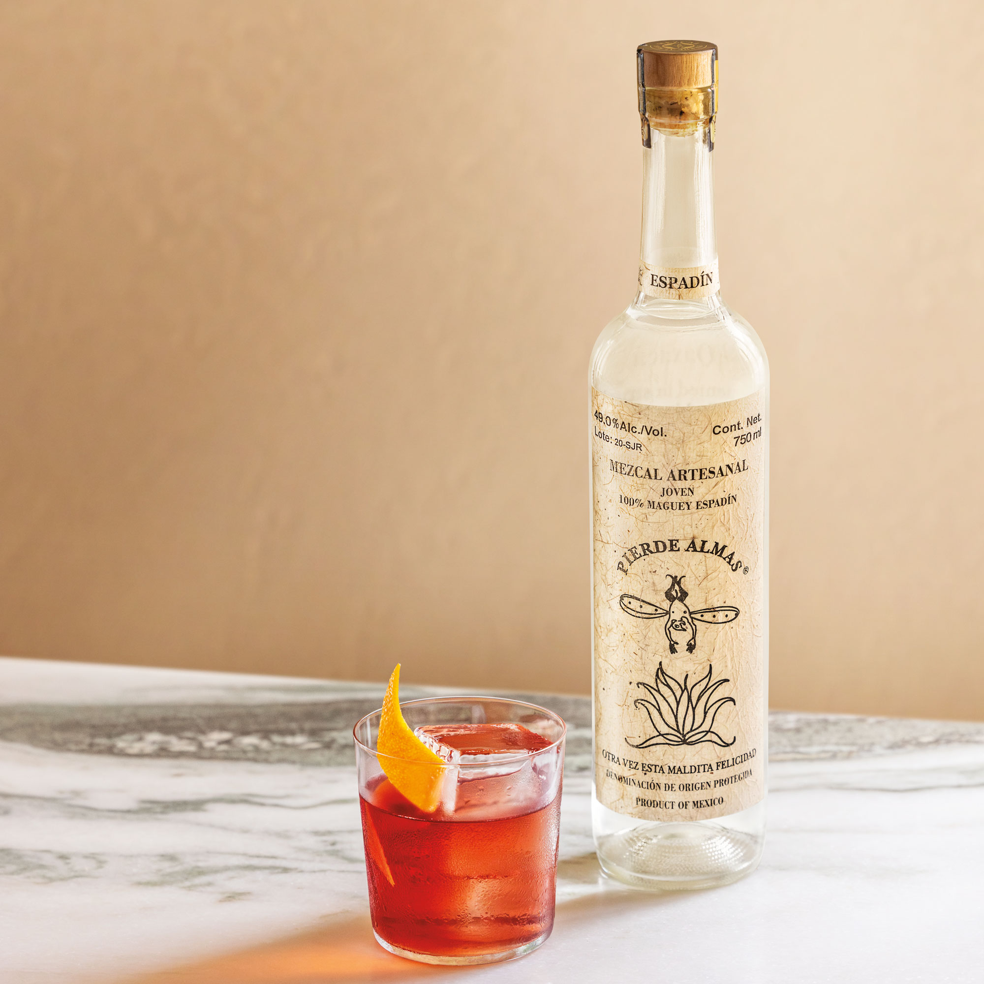 A bottle of Pierde Almas mezcal sits on a marble surface next to a glass of cocktail garnished with an orange peel.