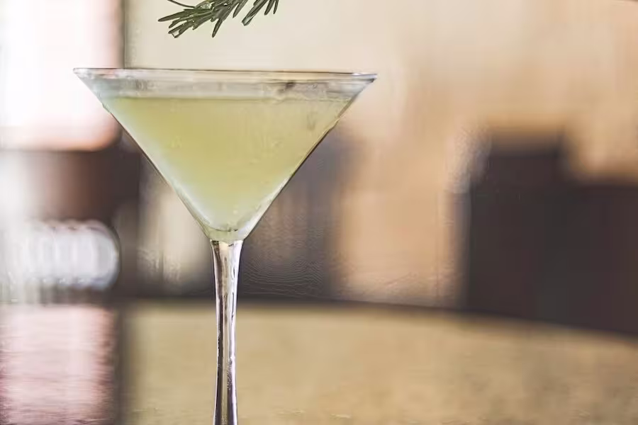 A yellow cocktail in a martini glass garnished with a sprig of rosemary, placed on a reflective surface with a blurred background.